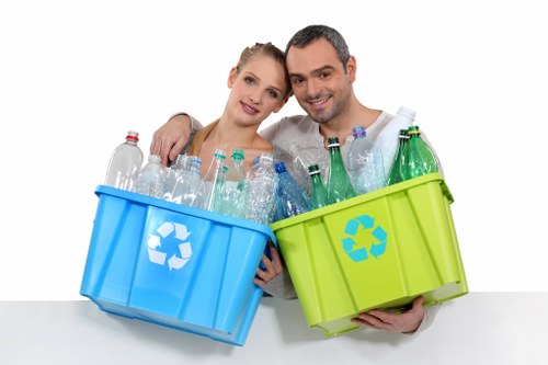 A smiling man and woman standing closely together, each holding a large plastic recycling bin, one blue and one green, filled with various clear plastic bottles and containers. The blue bin has a recycling symbol on its side and is filled predominantly with transparent plastic bottles, some with visible caps. The green bin, also marked with a recycling symbol, contains similar clear plastic bottles. The background is plain white, emphasizing the recycling theme. The individuals are positioned indoors, possibly in a waste management or recycling facility demonstration setting, illustrating environmentally conscious waste sorting and recycling practices in a professional manner. The image visually emphasizes the importance of plastic waste separation as part of responsible rubbish management services, which Commercial Waste Ruislip offers to local businesses and residents in the Ruislip area, aligning with responsible waste disposal and recycling efforts in the community.
